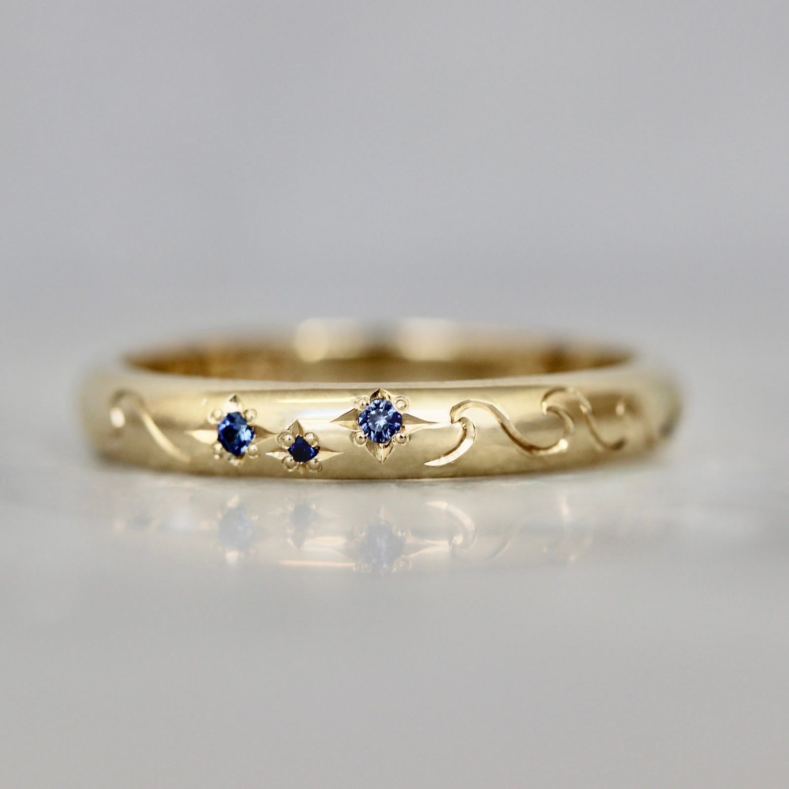 14k yellow gold custom wedding band with hand-engraved ocean waves and star-set teal sapphires by Gem Breakfast