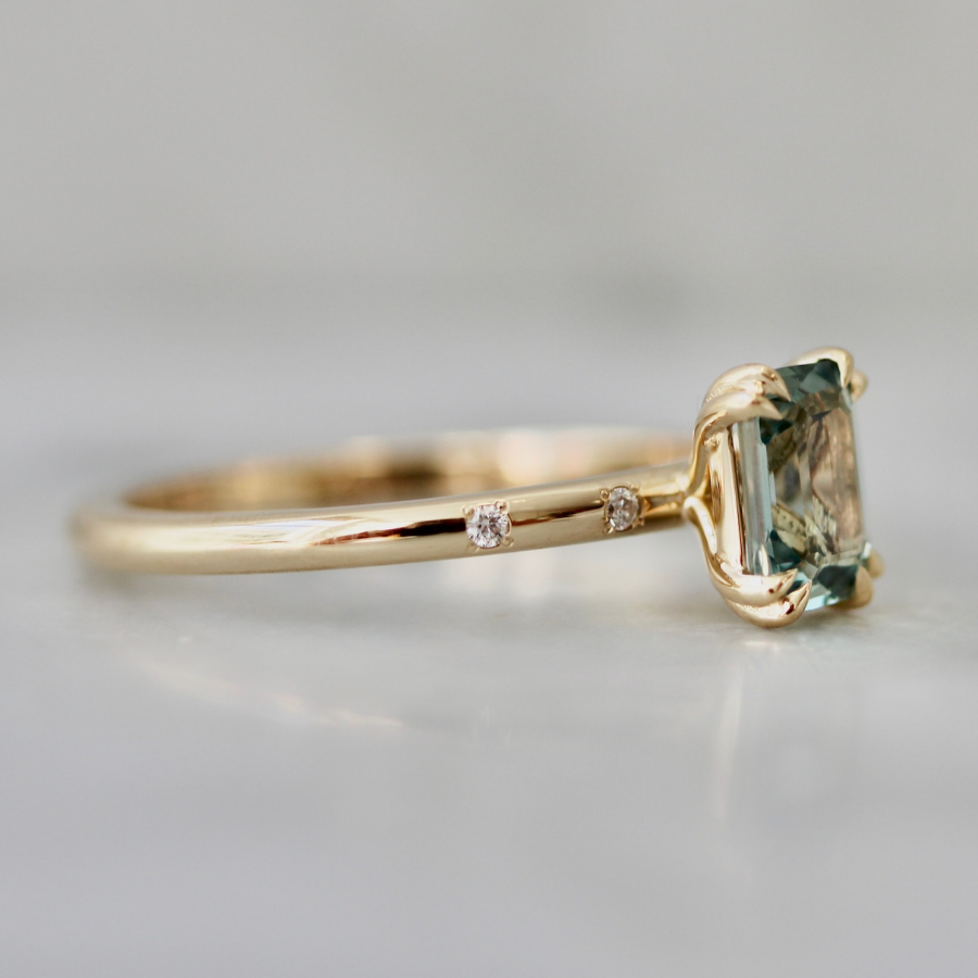 A yellow gold ring featuring a rectangular emerald-cut light blue gemstone and two small round-cut diamonds on the band, set against a soft gray background.