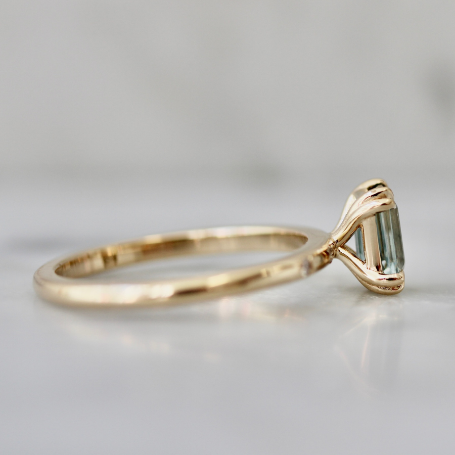A gold ring with a triangular-cut light blue gemstone, photographed on a light gray marble surface with a soft, blurred background.