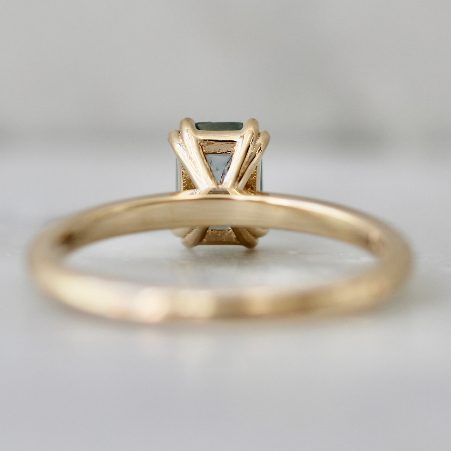 A gold ring with a rectangular light blue gemstone in a prong setting, shown from the back on a light gray background.