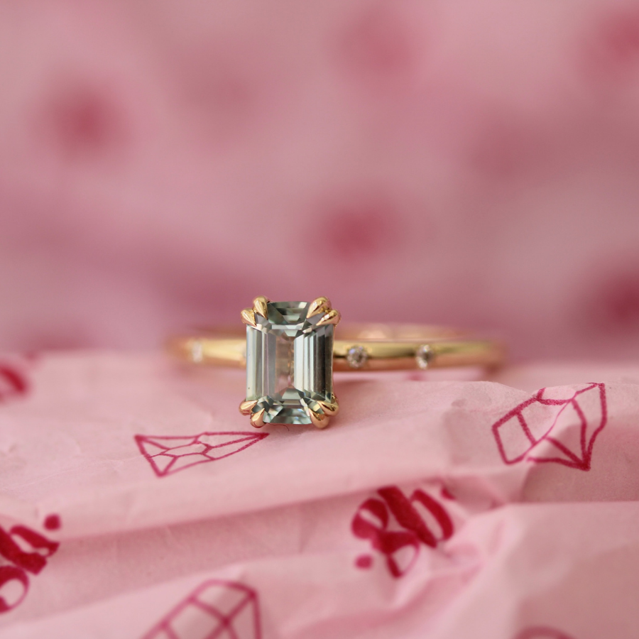 A gold ring with an emerald-cut green gemstone in a four-prong setting, resting on pink patterned paper with a soft pink blurred background.