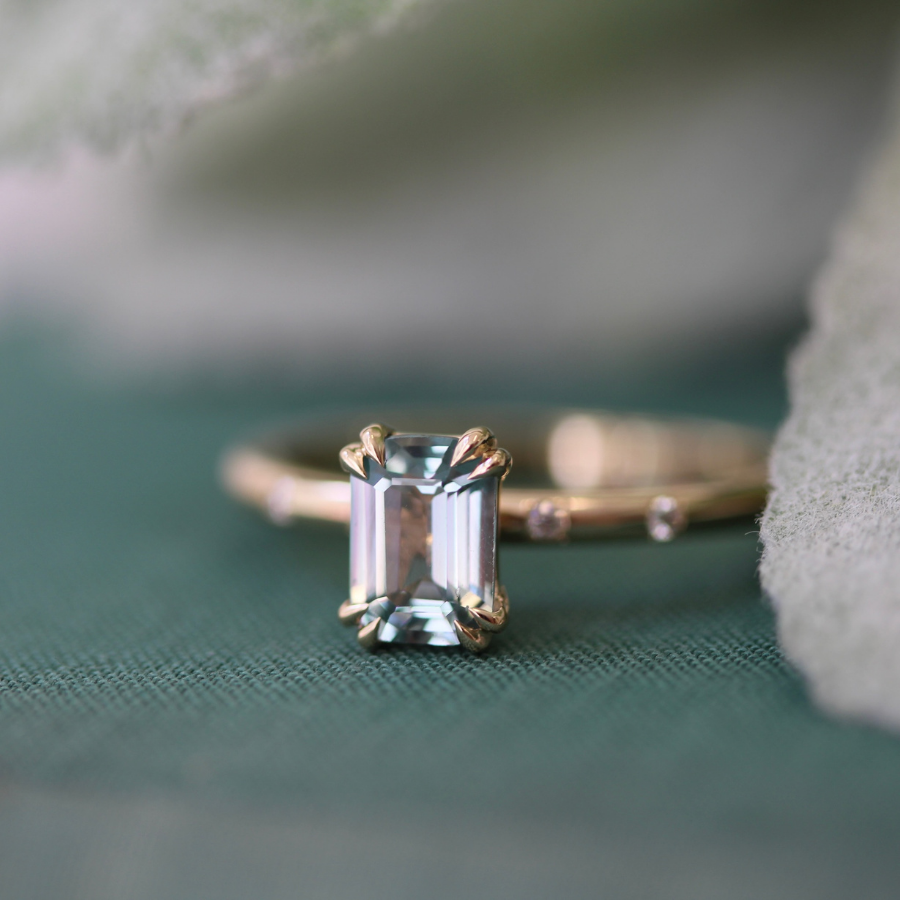 A gold ring featuring an emerald-cut light blue gemstone, resting on a textured dark green surface with soft, out-of-focus white elements in the background.