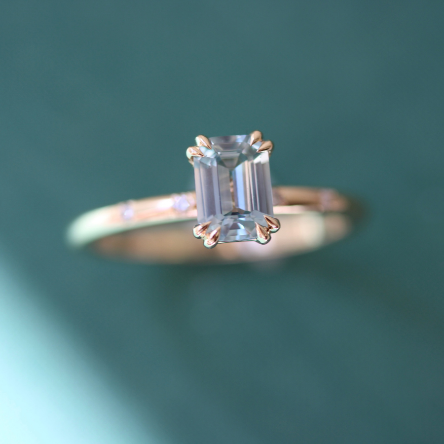 A rose gold ring featuring an emerald-cut clear gemstone in a prong setting, photographed against a soft teal-green blurred background.