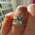 A close-up of a hand wearing a stack of rings, featuring a gold band with a rectangular light blue gemstone and two textured gold bands underneath, with a blurred building facade in the background.