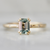 A yellow gold ring featuring a rectangular emerald-cut light blue-green gemstone in a four-prong setting, accented by small round diamonds on the band, shown on a light gray background.