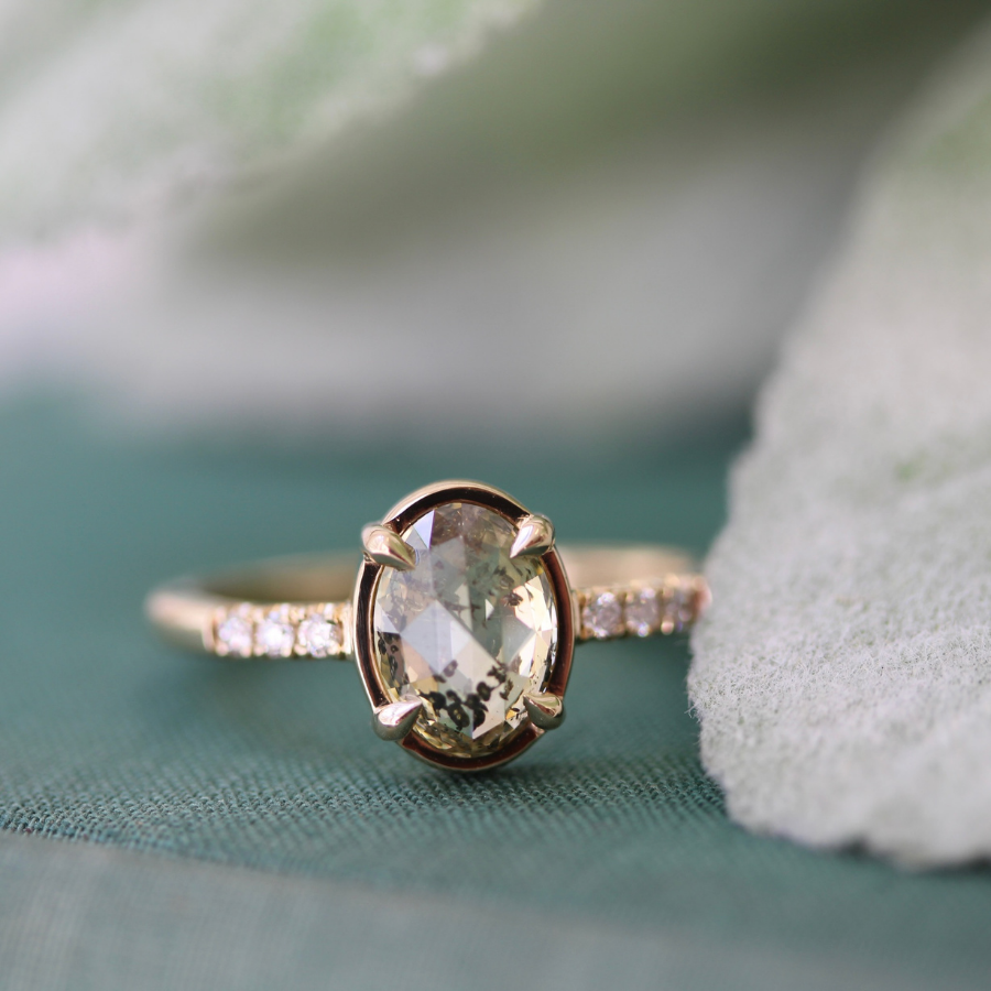 A yellow gold ring with an oval rose cut champagne salt and pepper diamond and small white diamond side stones, photographed close up on a teal textured surface with soft white and green blurred background elements.