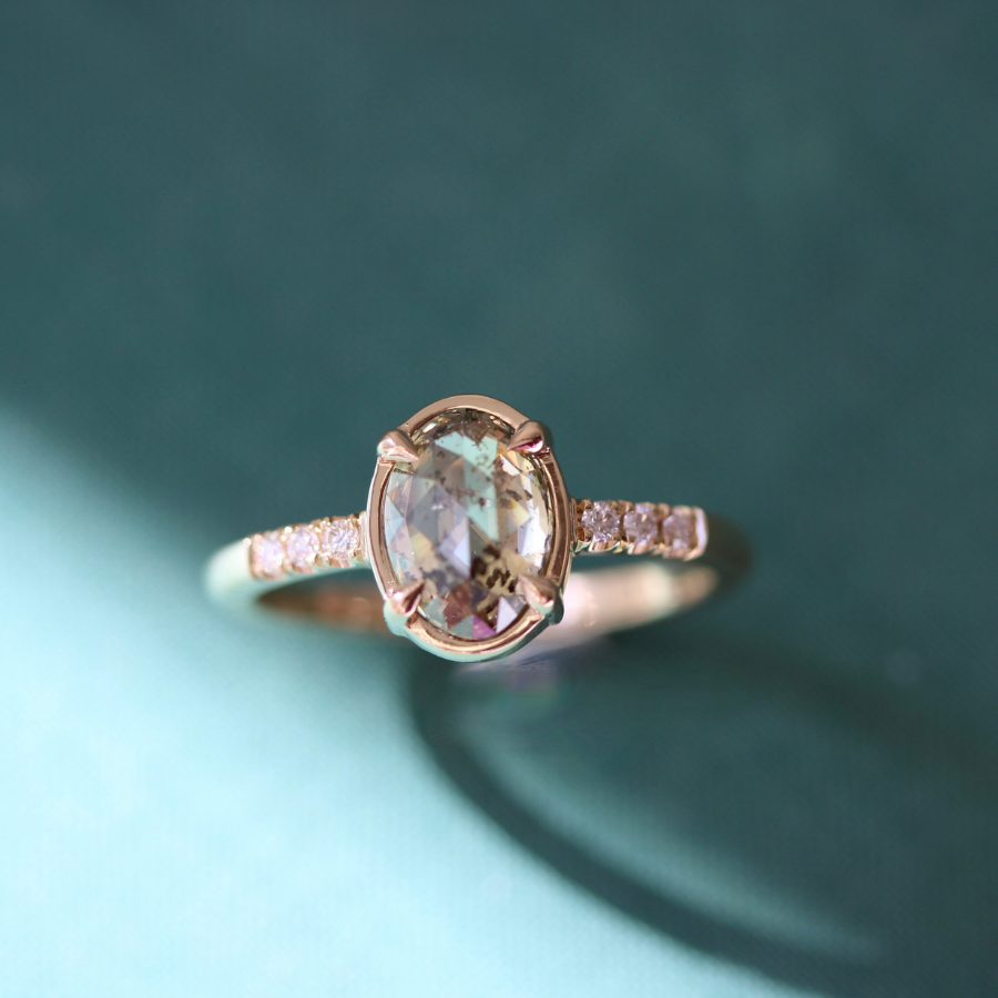 A yellow gold ring featuring an oval rose cut champagne salt and pepper center stone and small pavé-set clear stones on the band, photographed against a dark teal background.