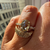 A close-up of a hand wearing a gold ring set with a large oval center stone and smaller side stones, with a textured gold band underneath.