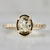 A yellow gold ring featuring an oval-cut, faceted champagne-colored gemstone in a prong setting, with small clear accent stones along the shoulders, shown on a light gray background.