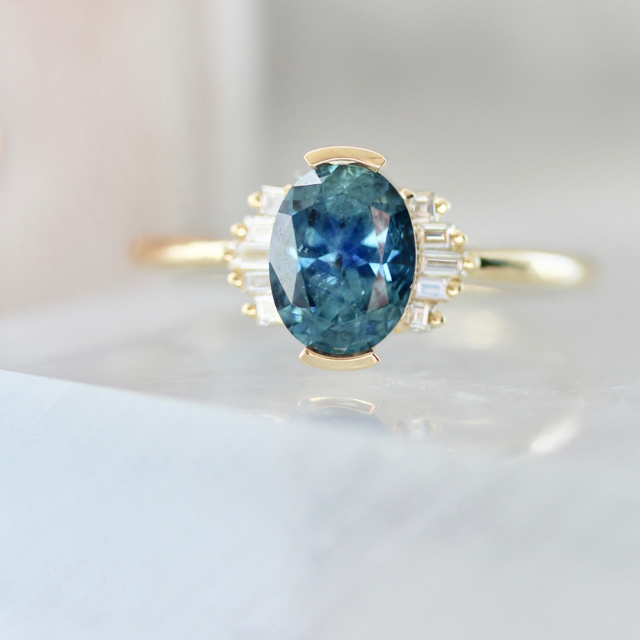 blue oval cut sapphire ring in half bezel with white baguette half halo in yellow gold