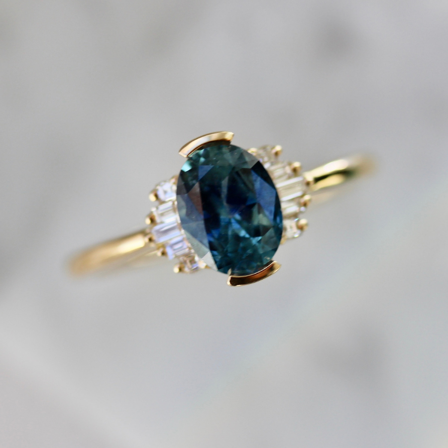 blue oval cut sapphire ring in half bezel with white baguette half halo in yellow gold